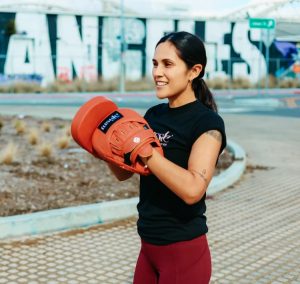 A woman wearing red boxing mitts and a black T-shirt smiles during an outdoor training session, demonstrating boxing mitts for general fitness use.