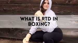 Female boxer in a hoodie and gloves kneeling against a wooden wall, with text overlay that reads “What Is RTD in Boxing?”—highlighting the main keyword.